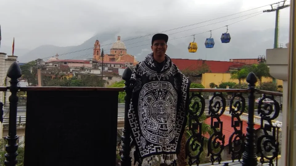 Yo, con un jorongo negro y blanco, en un balcón con barandal de herrería; atrás se ven un teleférico, una iglesia y montañas con neblina.